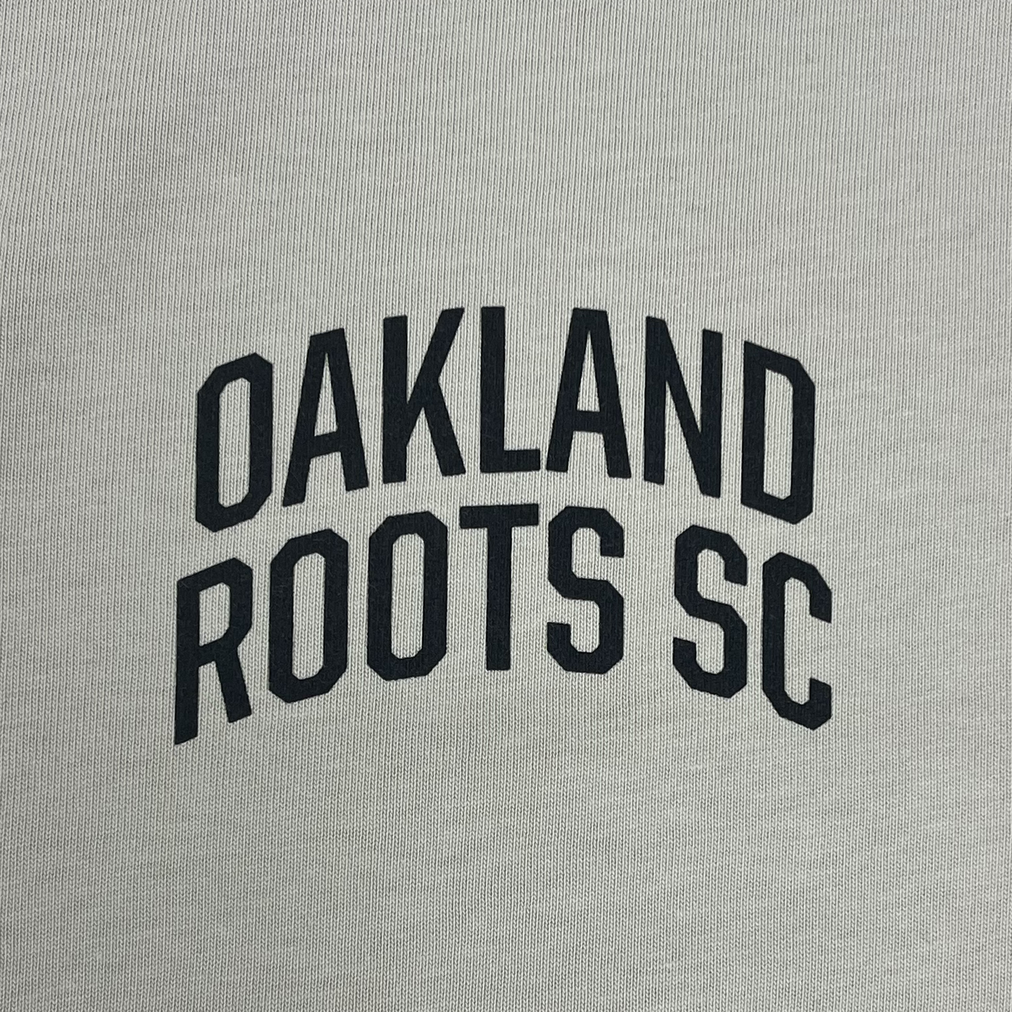 Close-up of black OAKLAND ROOTS SC wordmark on the left chest of a bone-colored button-up baseball jersey.