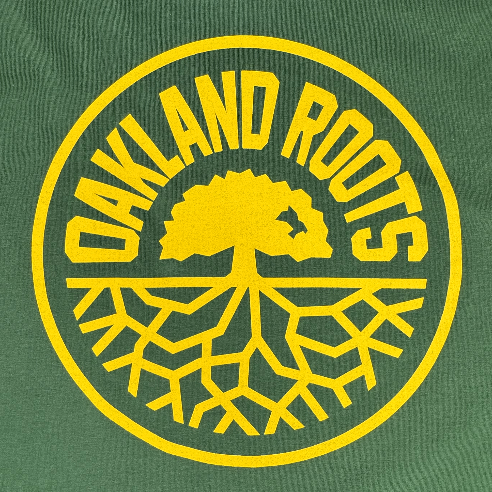 The Oakland Roots SC Classic 2.0 LS Tee by Oakland Roots SC features a circular logo with "OAKLAND ROOTS" in yellow encircling a tree with branches and roots on a green background, symbolizing community ties and social impact.