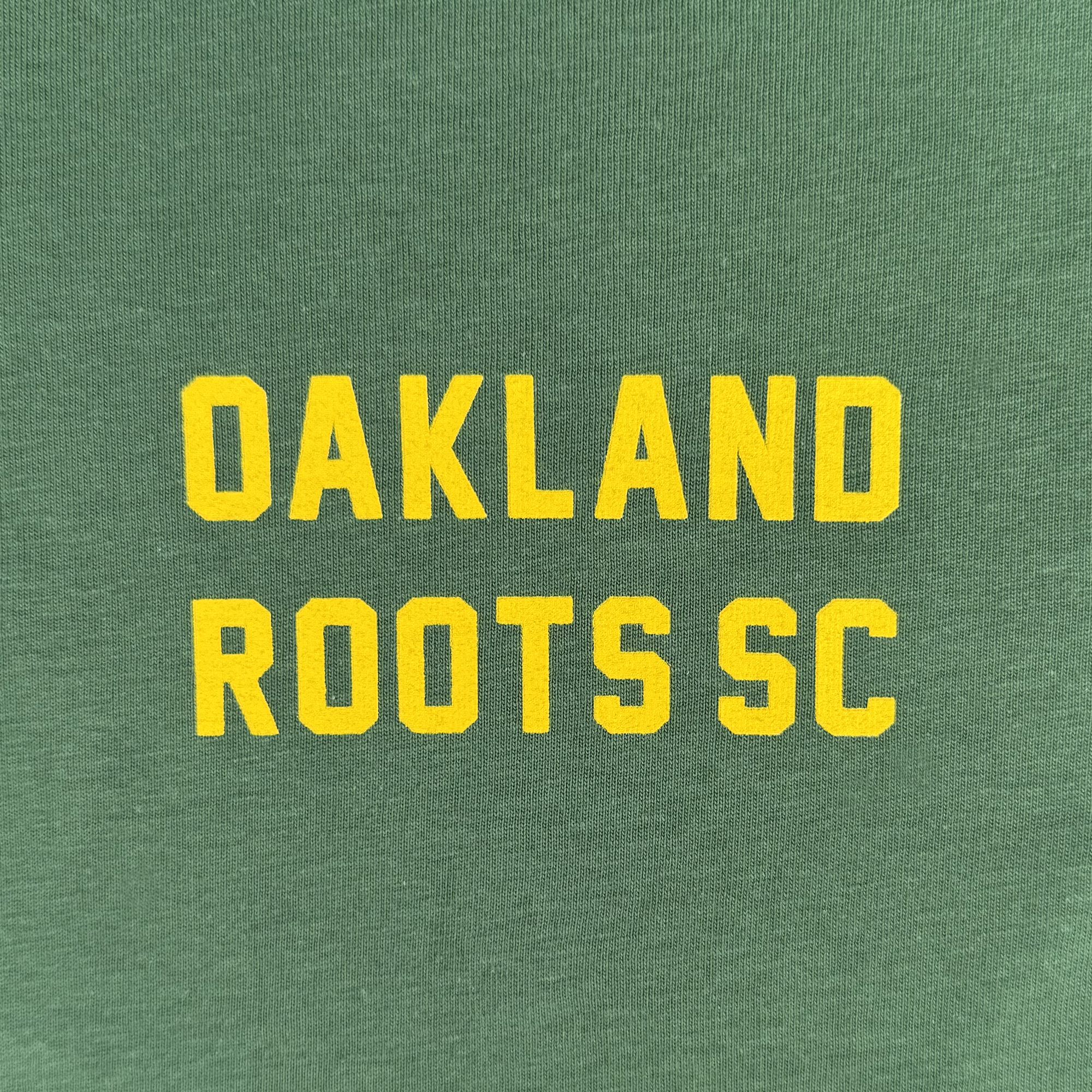 The Oakland Roots SC Classic 2.0 LS Tee features a green fabric background with bold, yellow, all-caps “OAKLAND ROOTS SC” text centered in blocky lettering, reflecting the athletic style of the Oakland Roots SC brand.