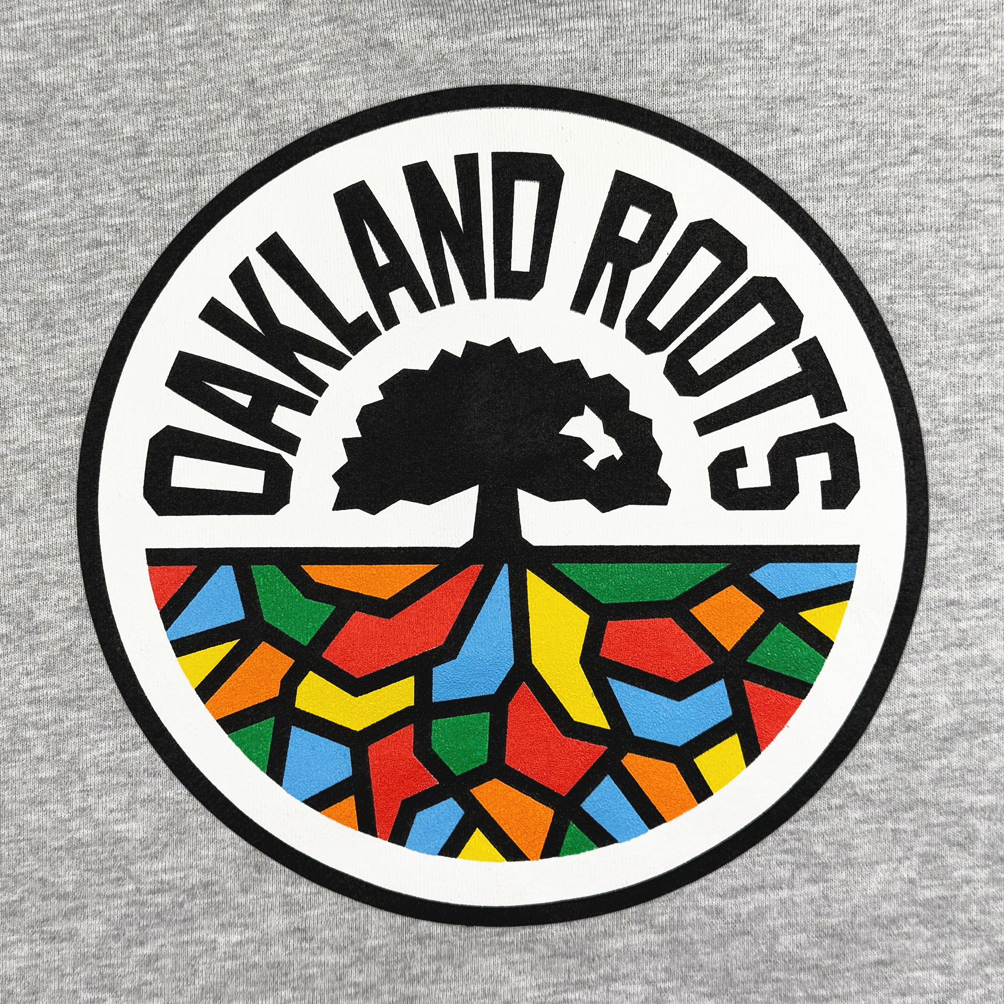 A circular logo on a gray background features the bold black text "Oakland Roots SC" from the Oakland Roots SC brand, representing a dynamic sports club. Beneath it, a stylized black tree stands atop a vibrant geometric mosaic with sections in red, yellow, green, orange, and blue, symbolizing its dedication to social good.