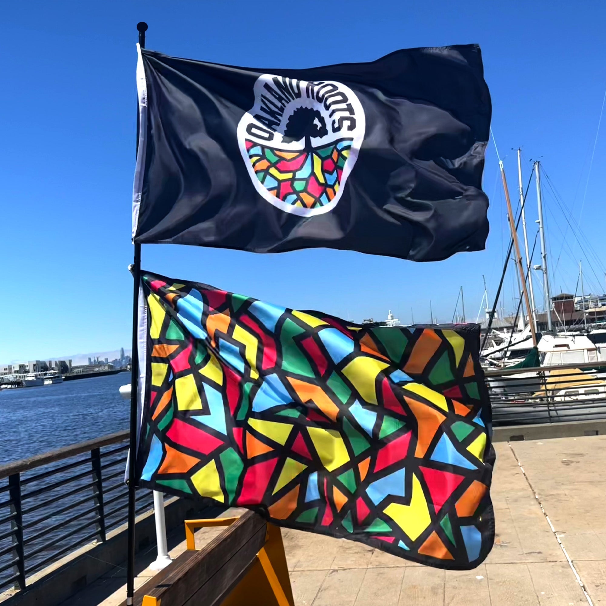 Outdoor photo of  Oakland Roots logo crest(top) and Oakland roots all over mosaic print (bottom) on pole 3' x 5' flag