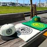 Image of Kelly green and Ecru t-shirt with Oakland Roots SC crest and NewEra snapback in Kelly and NewEra fitted hat in chrome photographed on outdoor picnic table.