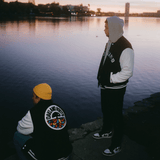 Two men are standing outdoors beside a lake, one side view to the camera, one with back to the camera, wearing Oaklandish varsity jackets.