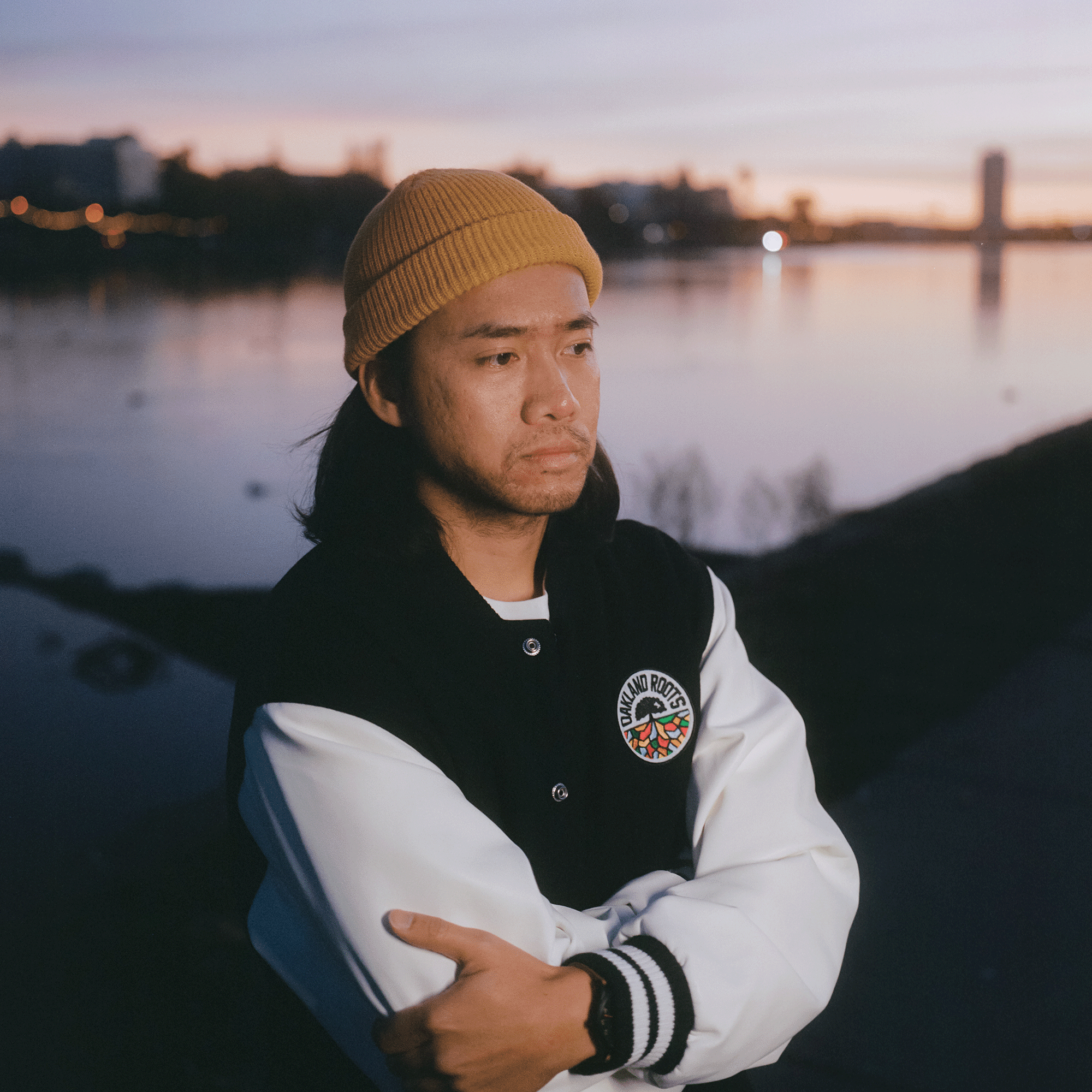 Man standing outside beside lake at sunset wearing black varsity jacket with white sleeves and black and white striped trim with full-color Oakland Roots logo on the left chest wearside.