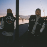 Two men, standing outdoors beside a lake, one facing the camera, one with back to the camera, wearing Oaklandish varsity jackets.